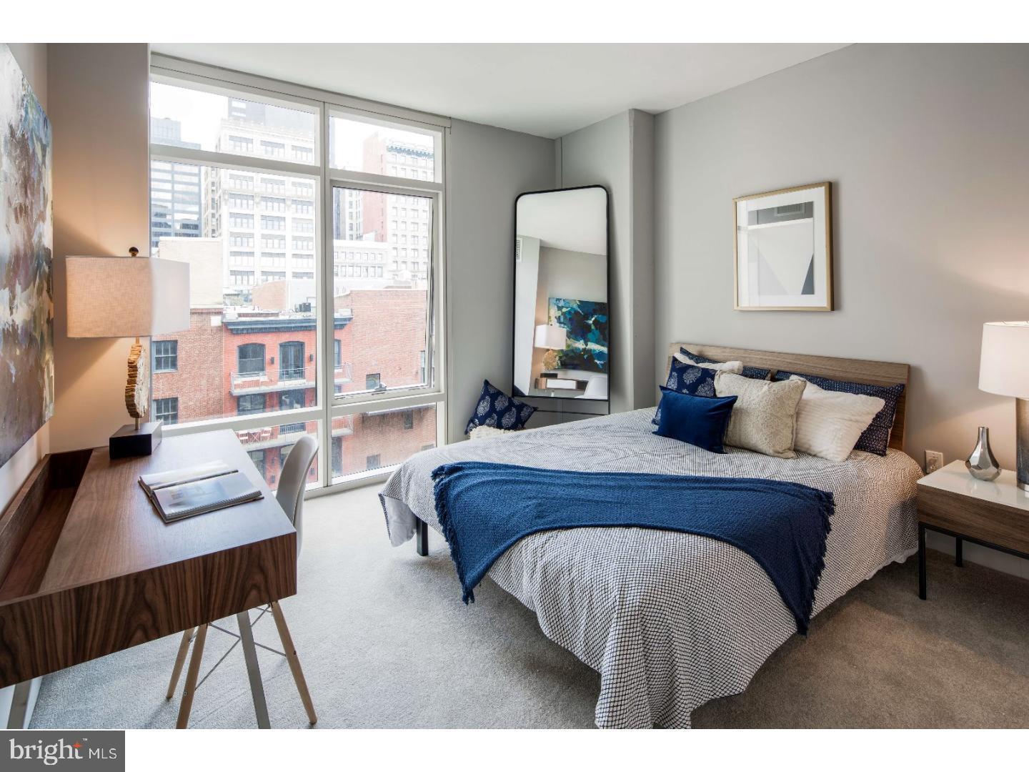 a bedroom with a bed a table and a large mirror next to a window