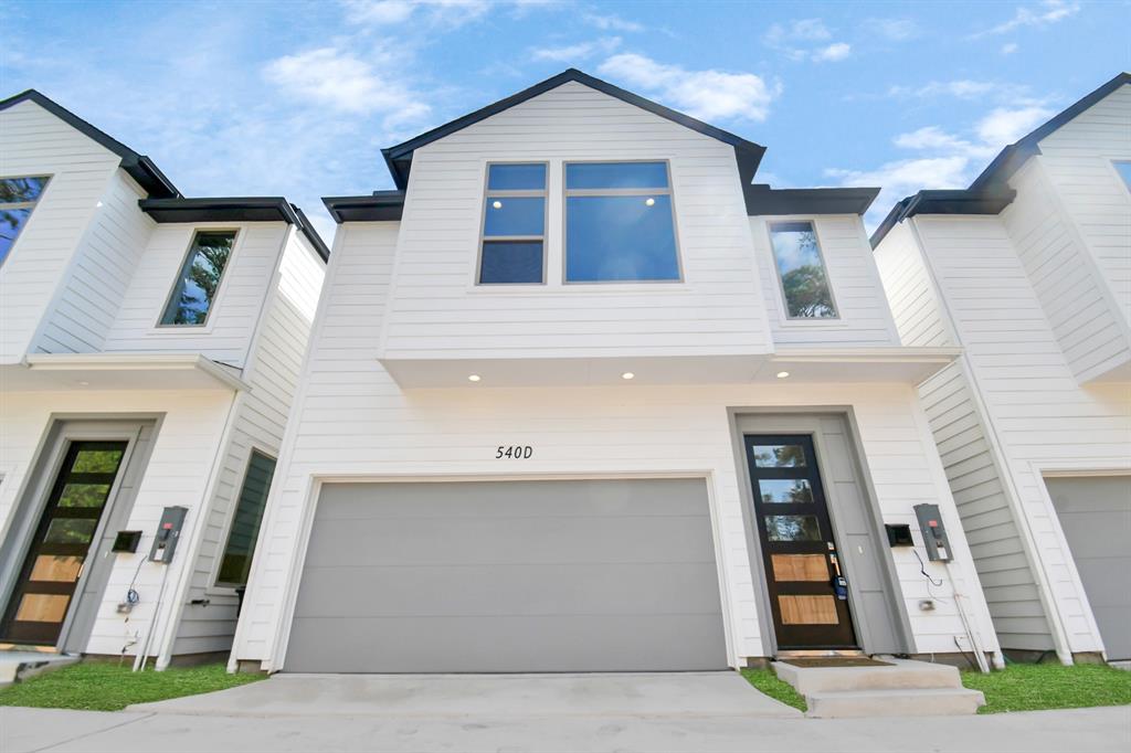 This aerial photo showcases a new townhouse development with gray roofs and white exteriors, nestled in a suburban setting with surrounding greenery and established trees. The neighborhood is adjacent to both commercial and residential areas, providing a blend of privacy and accessibility.