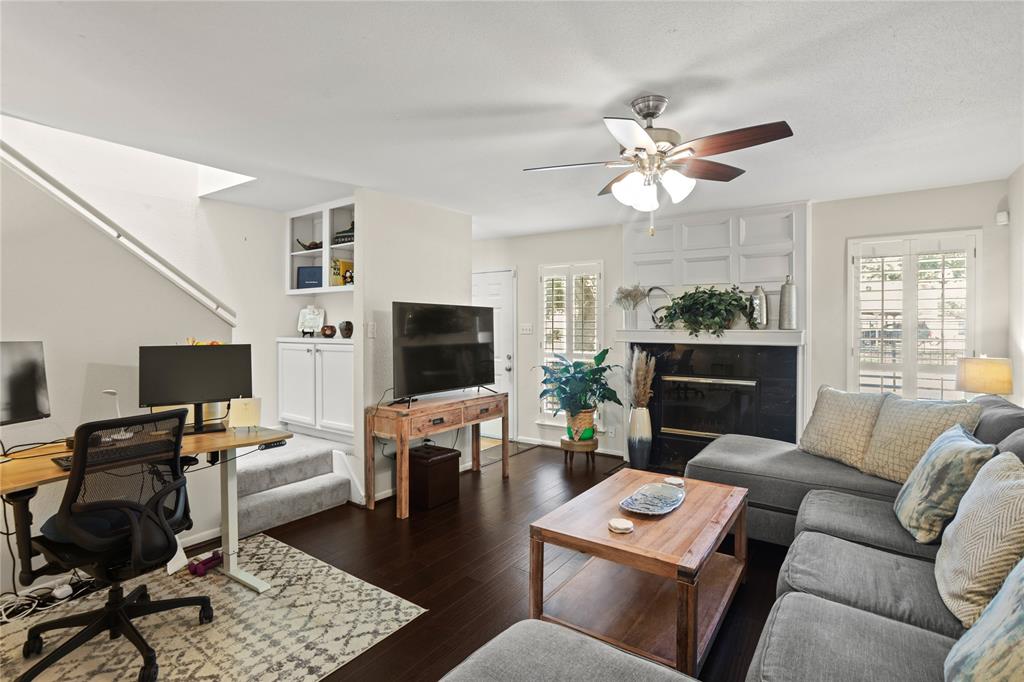 This is a cozy and well-lit living space featuring a comfortable sectional sofa, a modern ceiling fan, a fireplace with built-in shelves, and a dedicated workspace. The room is completed with engineered hardwood flooring and plantation shutters, offering a blend of functionality and comfort.