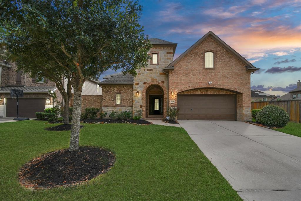 Charming single-family home featuring brick facade, two-story design, attached two-car garage, manicured lawn with mature tree, and a picturesque sunset sky.