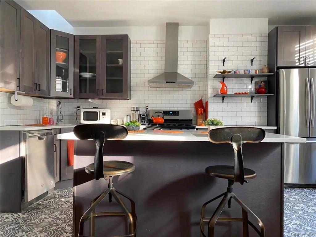 Kitchen featuring tasteful backsplash, appliances with stainless steel finishes, dark brown cabinets, a kitchen breakfast bar, and wall chimney exhaust hood