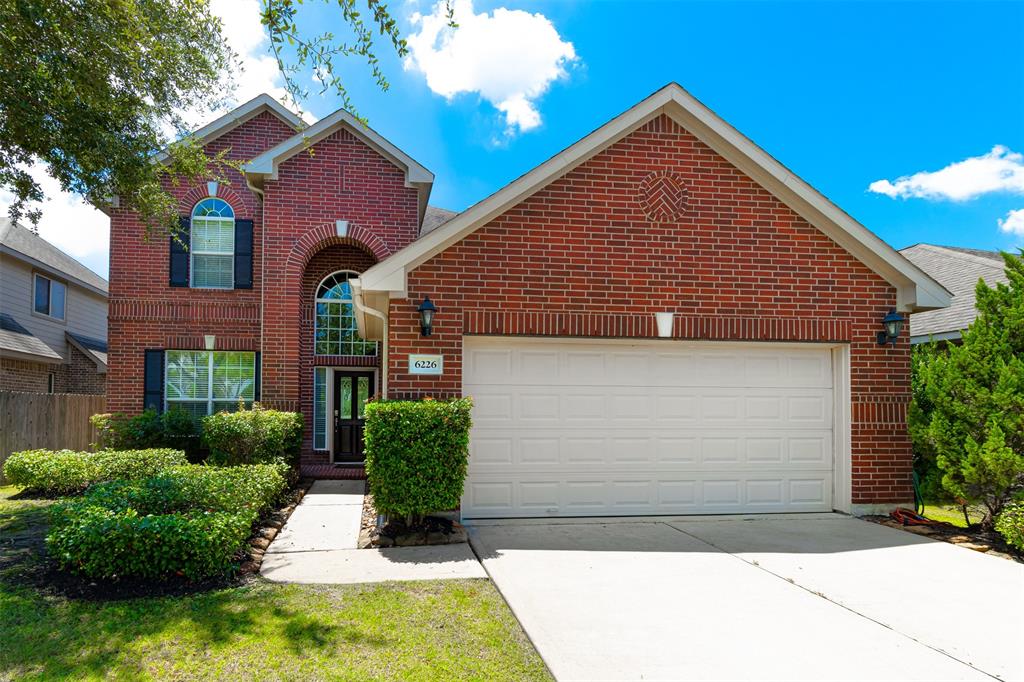 A two-story brick house with a white garage door, arched entryway, and manicured bushes under a clear blue sky.