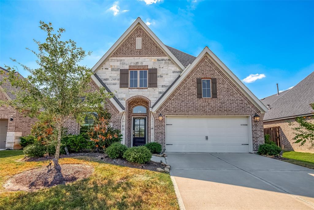 This is a spacious single-family home featuring a brick facade with a high-pitched roof, an arched entrance, and a two-car garage. The property includes well-maintained landscaping and a mature tree, offering a welcoming suburban curb appeal.