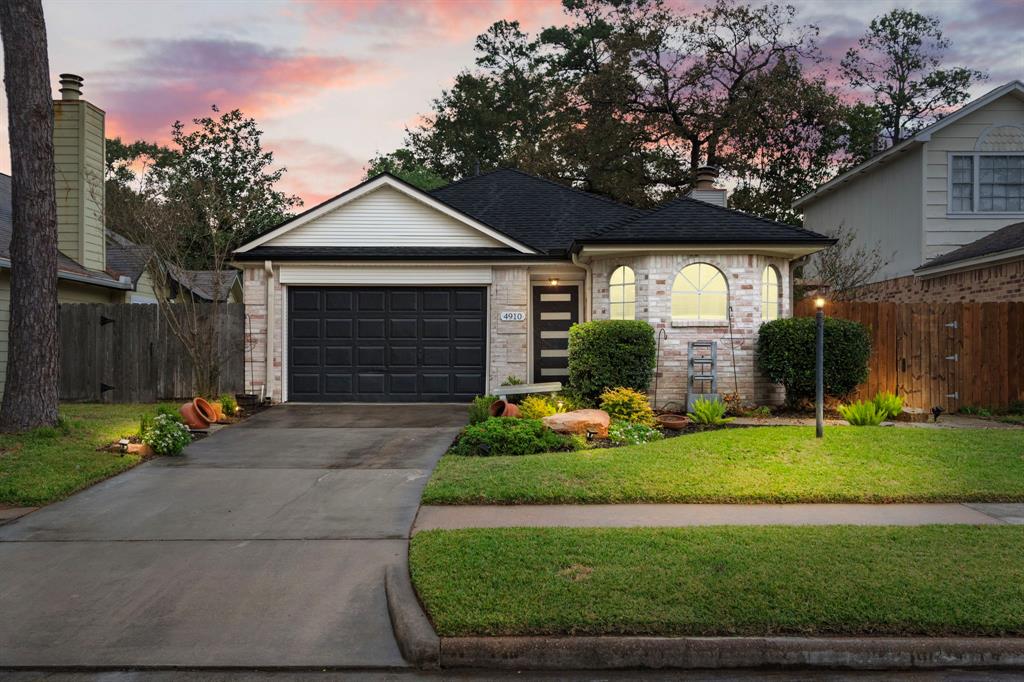 Clean, well-maintained front yard. It has a brick exterior, a black garage door, and a modern front door. The driveway leads to the garage, and the lawn is edged with landscaping, including a variety of plants and  decorative boulders.