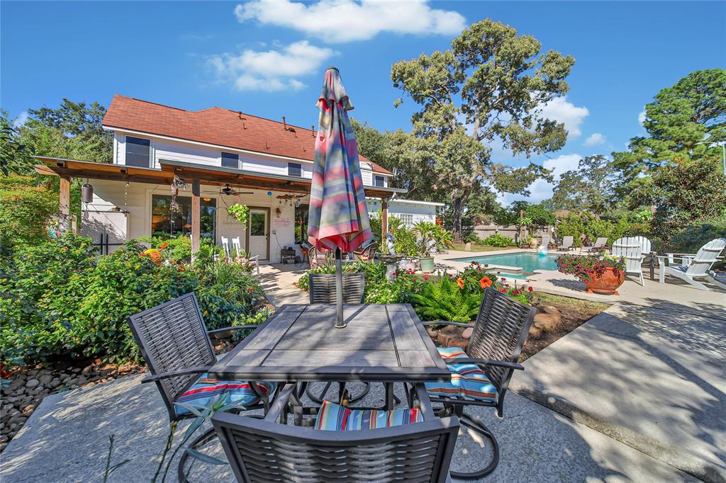 The well-maintained space includes an in-ground pool surrounded by lush landscaping and mature trees, offering a private and tranquil setting. The two-story home in the background has a classic design with a covered porch.