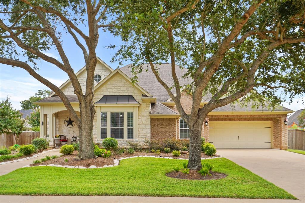 This is a single-story home with an attractive stone and brick elevation, a large two-car garage, and a neatly landscaped yard with mature trees. The home features a traditional design with a welcoming covered front patio and entrance.