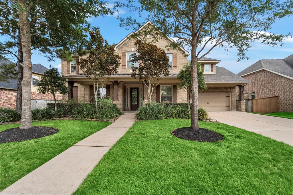 Beautiful two-story brick home with a manicured front yard, mature trees, and an attached three-car tandem garage. The house has a welcoming covered front entrance.