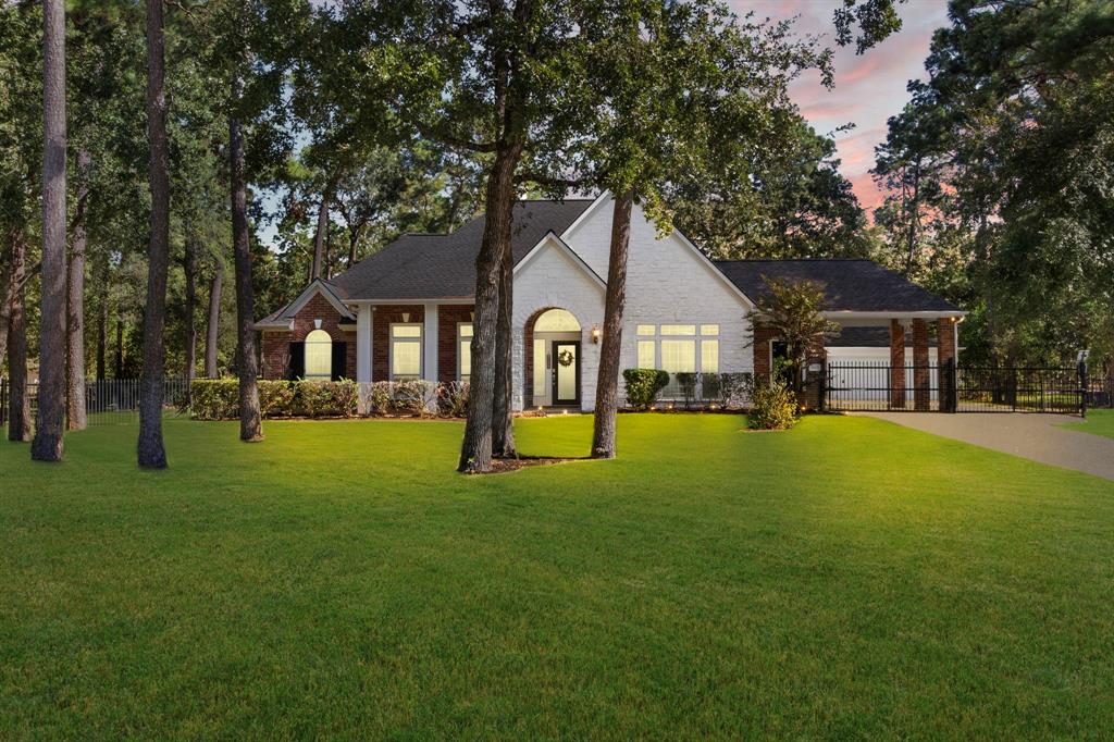 This is a single-story home with a brick facade, large windows, and a three-car garage, nestled amongst mature trees on a well-manicured lawn, featuring a gated driveway for added privacy.