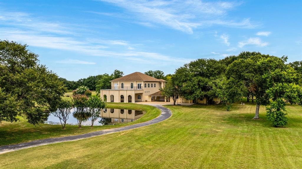 Spacious two-story home with a stucco exterior, featuring a sweeping driveway and surrounded by lush greenery. It overlooks a tranquil pond, offering picturesque views and a serene living environment. The 3521 sq ft guest house is equally impressive.