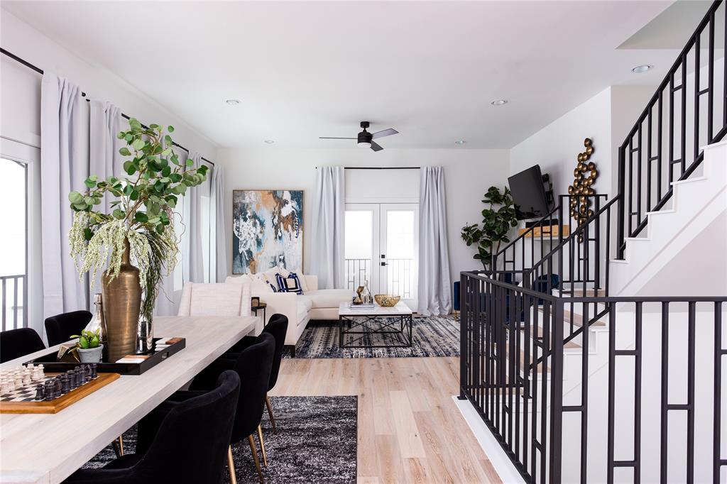 This modern open-plan living space features a combined dining and lounge area with light hardwood floors, elegant decor, and tasteful artwork. Ample natural light flows through large windows dressed with soft curtains, enhancing the inviting ambiance. The staircase with black railings adds a contemporary touch, leading to the home's upper level.