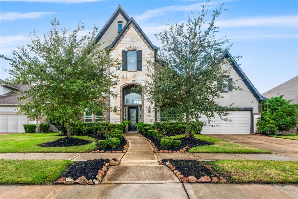 This is a spacious two-story stone and stucco home with a steeply pitched roof, featuring an arched entryway, a double garage, and well-manicured landscaping with mature trees.