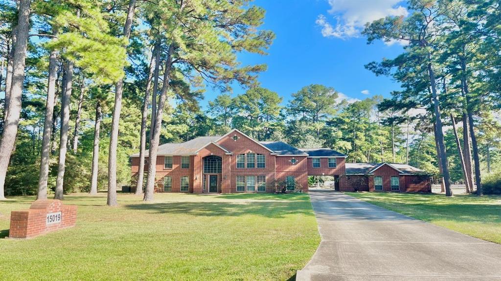 This is a two-story brick home nestled among tall pine trees, featuring a large yard, an attached garage, and an expansive driveway.