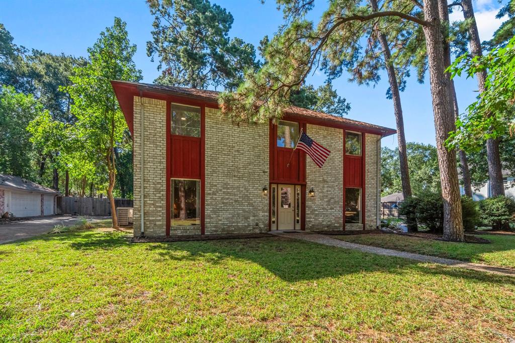 Charming two-story brick home surrounded by mature trees in a serene neighborhood. Features include a prominent red door, spacious front yard, and a welcoming American flag.