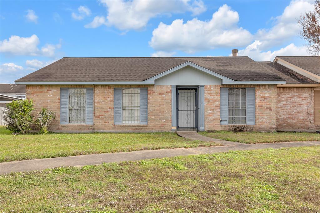 This is a single-story brick home with a traditional design. It features a central front door flanked by two large windows with blue shutters. The property has a modest front yard and a paved walkway leading to the entrance.