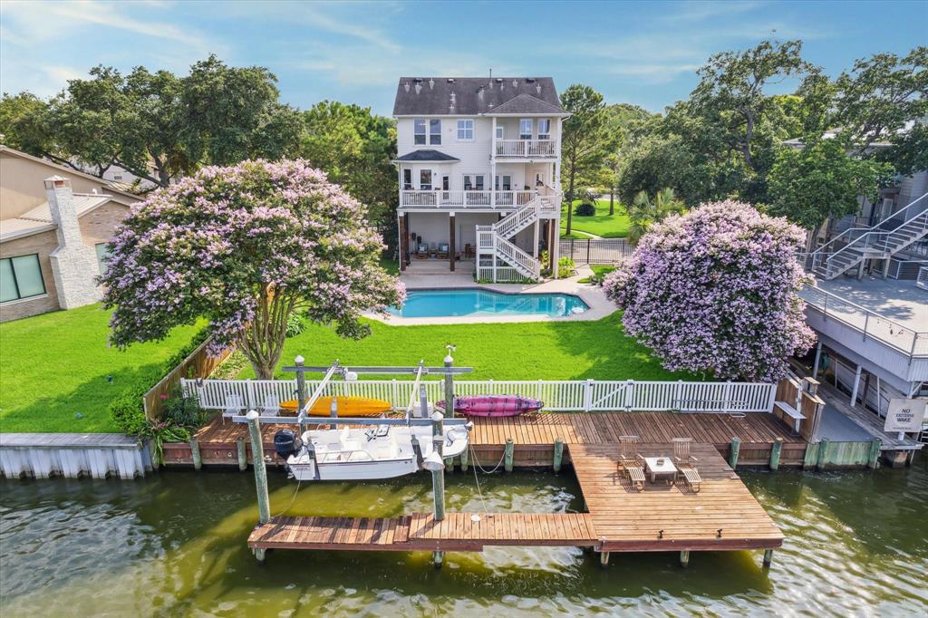 The expansive backyard offers a private waterfront paradise, featuring a large pool surrounded by lush greenery. The custom boat dock provides access to the water for boating and kayaking, with serene views of the bay and beautiful flowering trees lining the property.