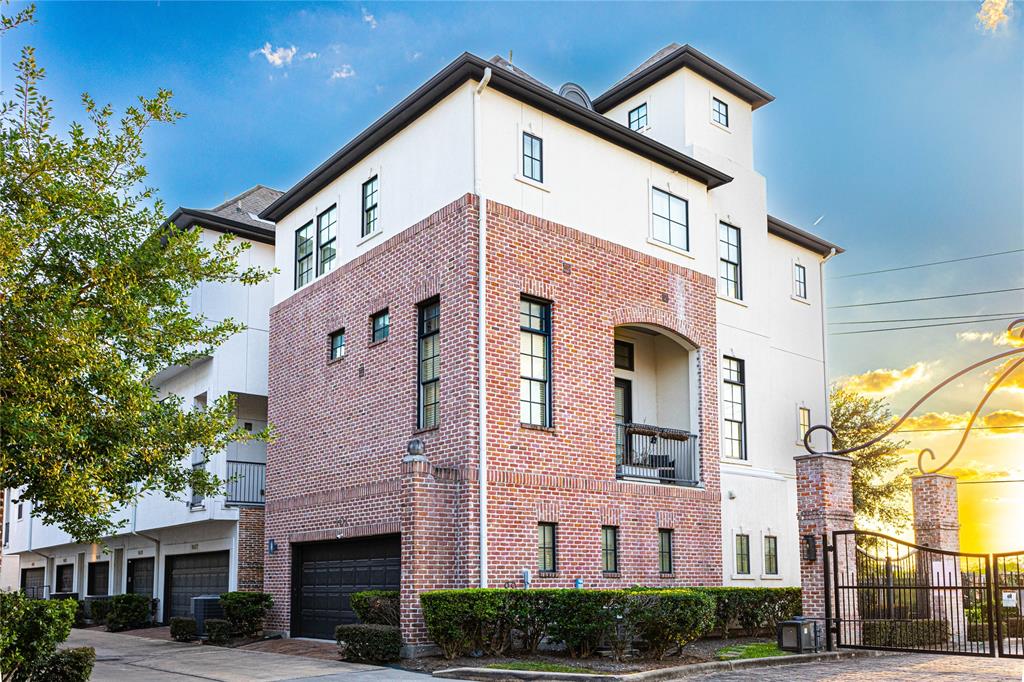 This is a modern three-story house with a white and brick facade, featuring large windows and a wrought iron fence. It's nestled in a well-maintained area with landscaping and mature trees.