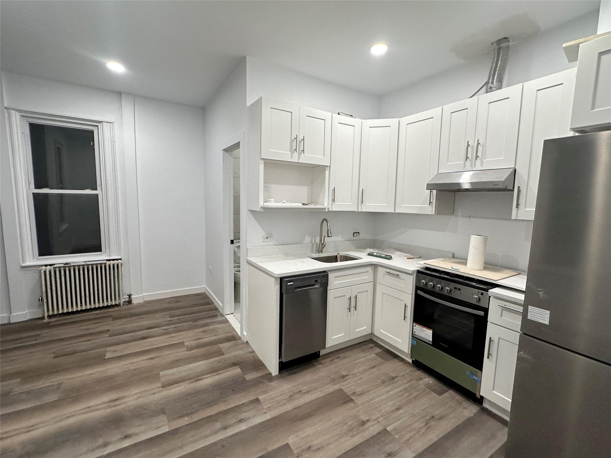 Kitchen with radiator, ventilation hood, stainless steel appliances, dark hardwood / wood-style floors, and white cabinetry