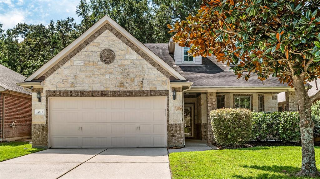 This is a single-story home featuring a stone and brick facade with a two-car garage. Landscaping includes mature trees and well-maintained shrubbery, contributing to its curb appeal.