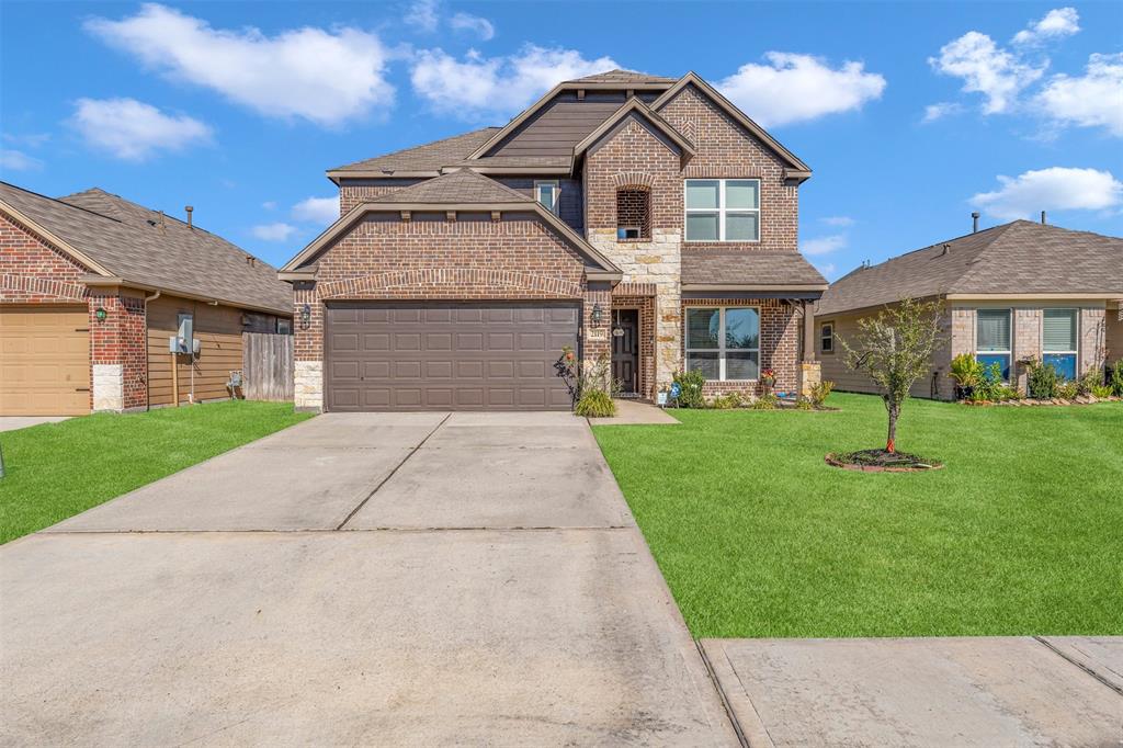 This is a two-story brick home featuring a two-car garage, manicured lawn, and a welcoming entryway. The design includes stone accents around the front door and a small, young tree in the front yard.