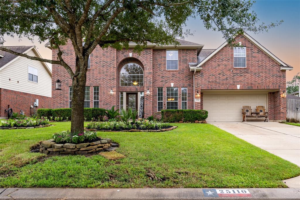 This is a two-story brick home featuring a large arched window, an attached two-car garage, and a well-manicured front lawn with a mature tree and landscaping.