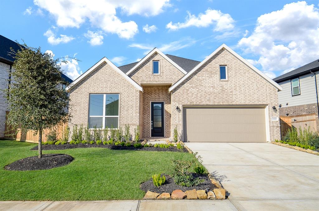 This is a modern single-story brick home with a gabled roof, featuring a spacious driveway and a two-car garage. The front yard is well-maintained with a small tree and neat landscaping.