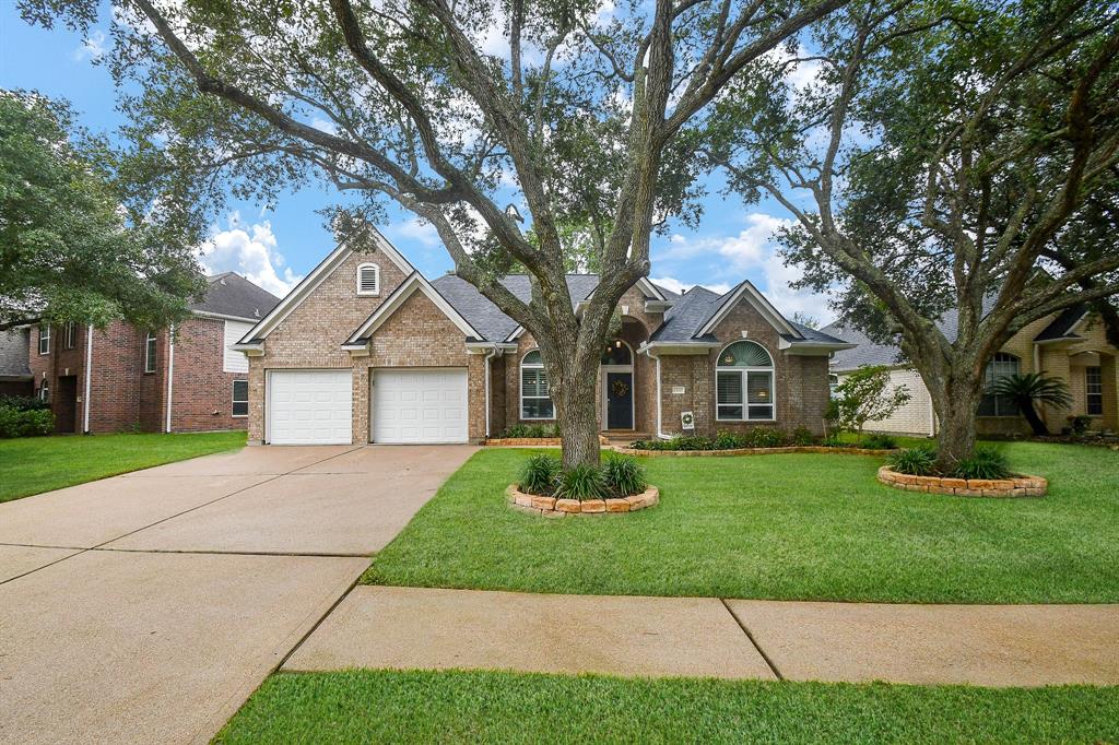 This is a single-story brick home with an attractive roofline and two-car garage. It features an arched front doorway and large windows, nestled under mature trees on a well-manicured lawn.