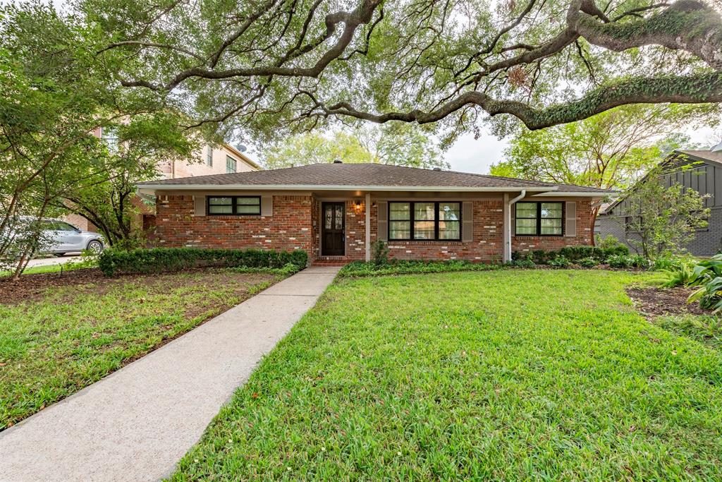 This is a single-story brick house with a well-maintained lawn, shaded by mature trees, and featuring a welcoming walkway leading to the front door. The home has large windows and a traditional design.