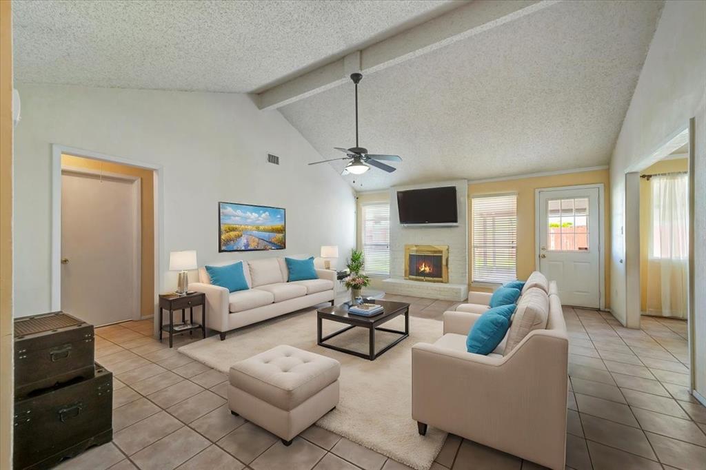 This is a bright and airy living room featuring vaulted ceilings with ceiling fans, a fireplace, tiled flooring, and natural light flowing in through windows and glass doors leading to an outdoor area. The space is furnished with a comfortable beige sofa set and a central ottoman.