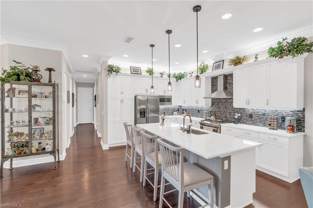 Kitchen with stainless steel appliances, sink, wall chimney range hood, decorative light fixtures, and a center island with sink