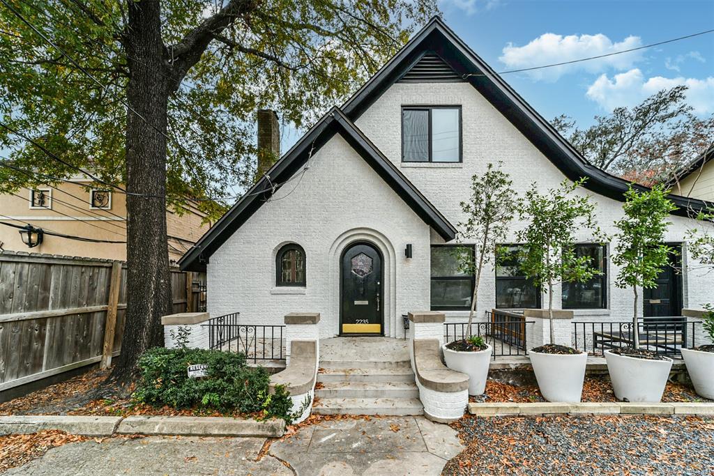 Charming two-story duplex with a classic gabled roof and white brick exterior. Features a welcoming arched doorway, large windows, and mature trees. The front yard includes tasteful landscaping with potted plants and a stone walkway.