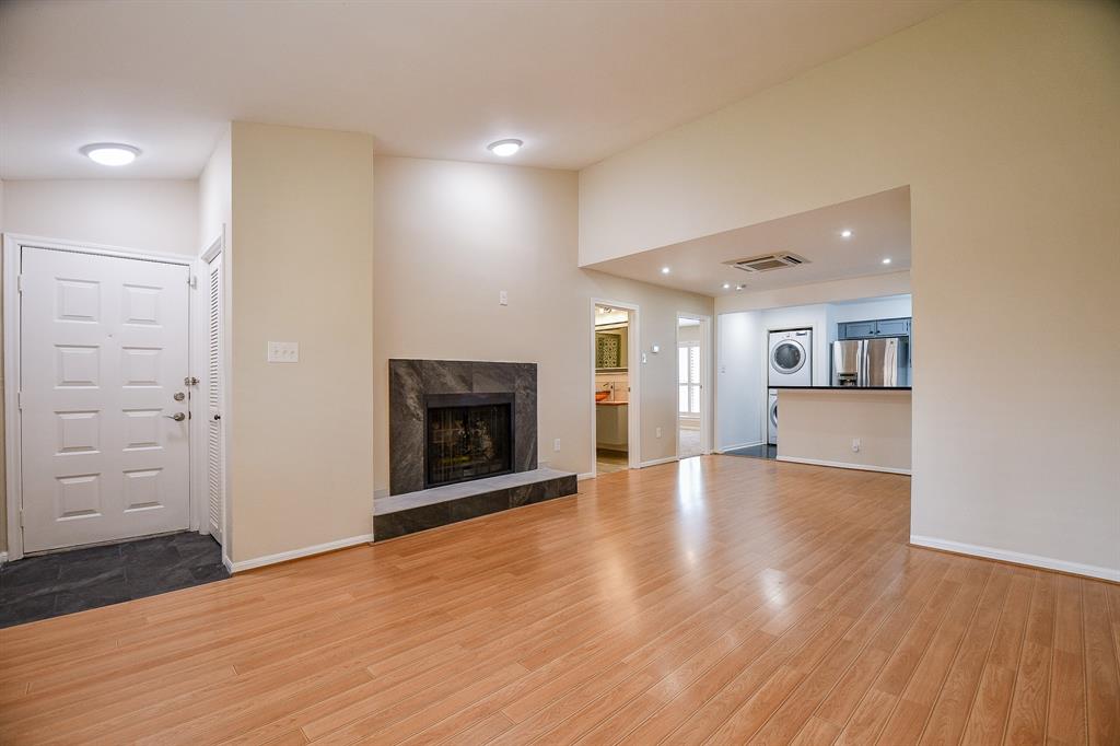 This is a spacious, well-lit living area featuring hardwood floors, a cozy fireplace, recessed lighting, and a view into a modern kitchen with stainless steel appliances. The entryway includes a convenient coat closet.