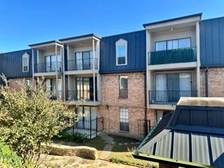 This photo shows a modern apartment building with three floors, featuring brick and dark paneling. Each unit has a balcony, and the surrounding area includes a small landscaped garden. The overall design is clean and contemporary.