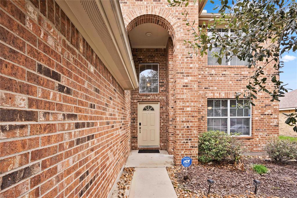 This is a two-story brick home featuring a welcoming front entrance with a tall arched entryway, a ground-level window, and mature landscaping. It has an existing security system, as indicated by the yard sign.