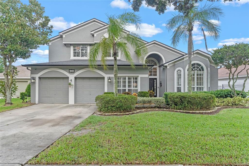 Exterior Front View: This image shows a two-story house with gray exterior paint, white trim, and a double garage. A neatly manicured lawn, landscaped with shrubs and palm trees, leads up to the arched entryway and large windows with white shutters. The driveway and sidewalk are visible, with trees adding shade in the foreground. The house has a clean, modern look with prominent gables and a welcoming design.