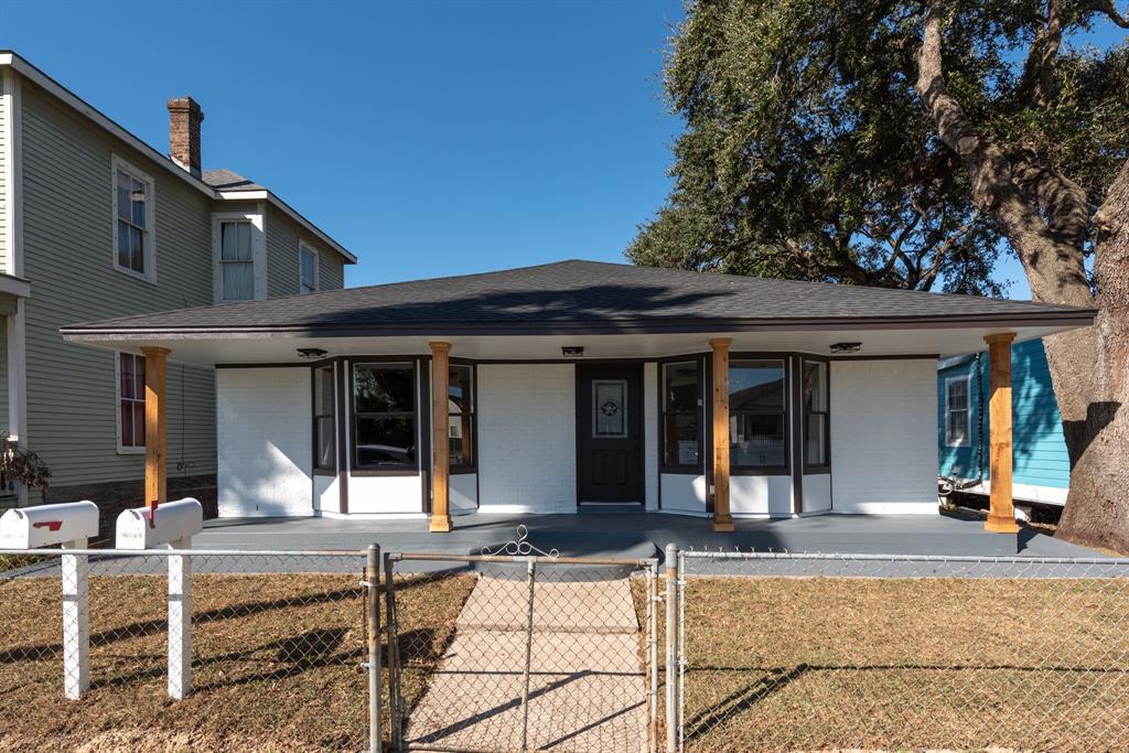 This is a single-story home with a large front porch, supported by wooden columns, and a fenced yard. The exterior is white with contrasting window trim, and there’s a mature tree to the right side, offering shade.