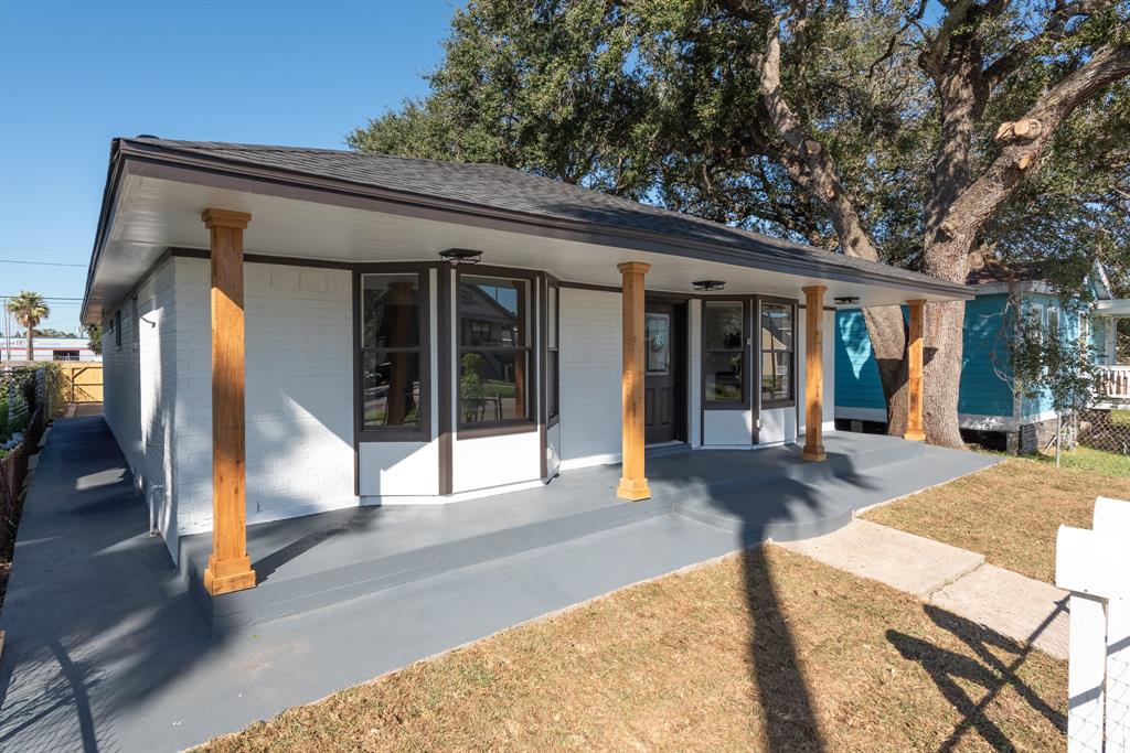 This is a single-story home featuring a freshly painted exterior with white walls and black trim, a covered front porch with wooden columns, large windows, and a spacious concrete patio area. The property includes mature trees and a fenced yard and easy access to the road.