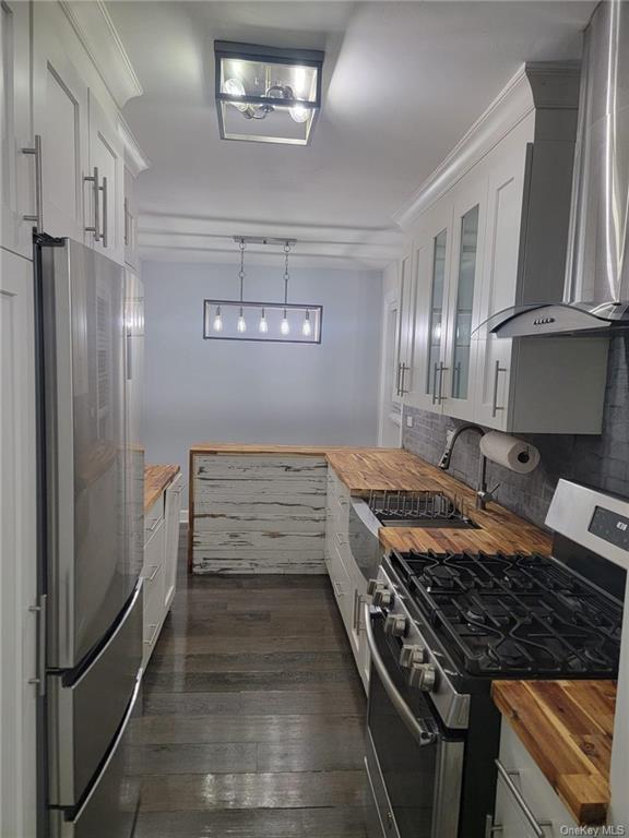 Kitchen featuring appliances with stainless steel finishes, white cabinetry, wood counters, and wall chimney range hood