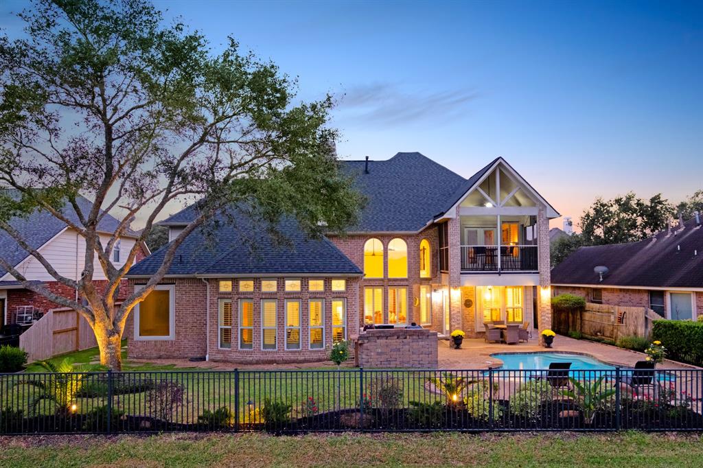 A spacious two-story brick home featuring large windows, a balcony, and an inviting backyard with a pool, all enclosed by a black wrought-iron fence. The property offers an elegant evening ambiance with warm interior lighting and tasteful landscape lighting.