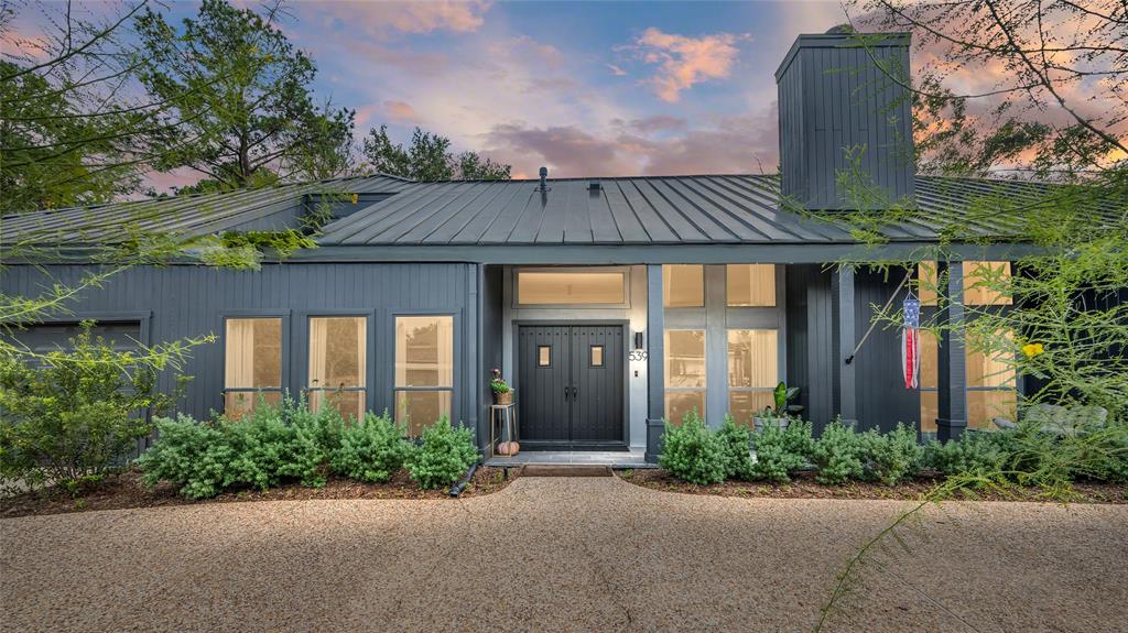 This modern home features a sleek gray exterior with a metal roof, large windows, and a welcoming front door. The house is surrounded by lush greenery and has a stylish, contemporary design.