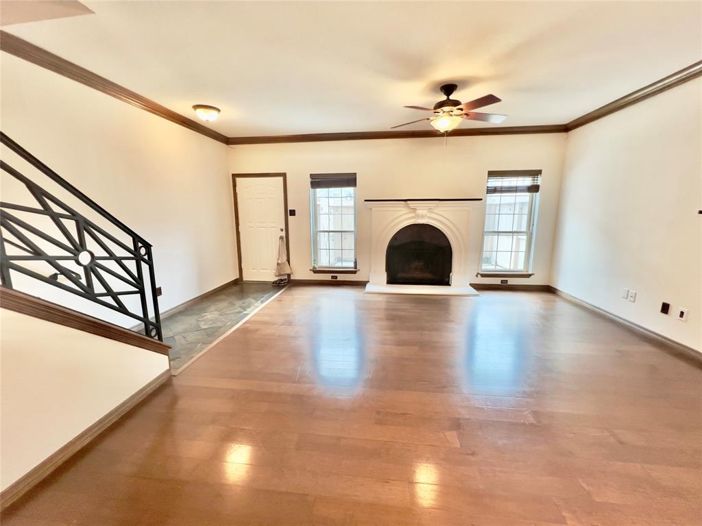 This photo features a spacious, well-lit living room with hardwood floors, an elegant fireplace, a ceiling fan, and a stylish staircase leading to the upper level. The room includes a door and two large windows that allow for natural light.