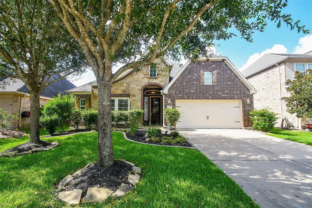 This is a charming single-family home featuring a brick facade, an arched entryway, a two-car garage, and mature landscaping with trees. The property offers a well-maintained lawn and a welcoming suburban feel.