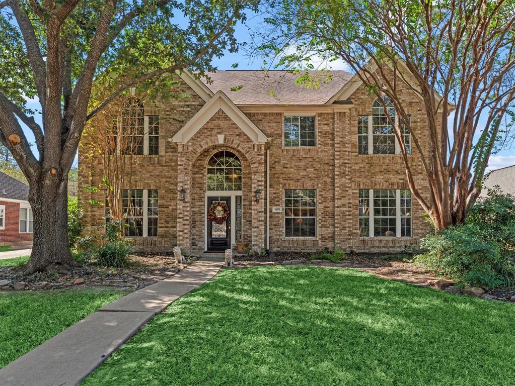 This charming two-story brick home features large windows and a welcoming arched entryway. It is surrounded by mature trees and lush landscaping, creating a beautiful and inviting curb appeal.