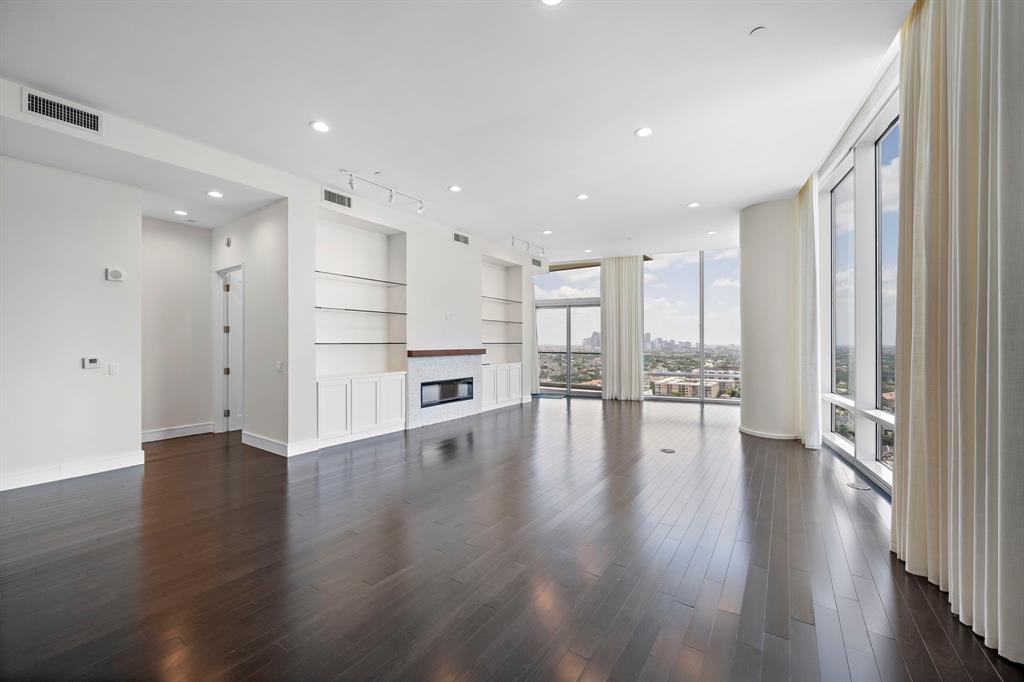 This is a spacious, living room with hardwood floors, floor to ceilings windows offering plenty of natural light, and a cityscape views. It features a built-in fireplace and shelving, high ceilings, electric shades & drapes with a clean, white interior palette that awaits personal touches.