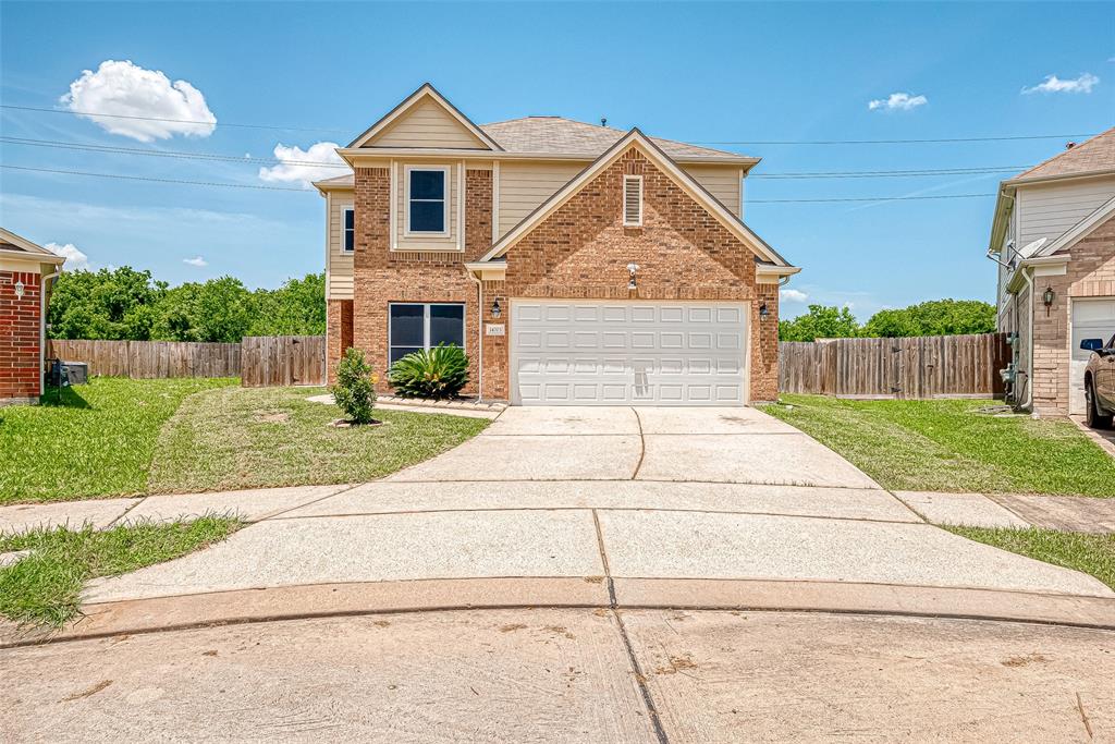 This is a two-story brick home featuring a single-car garage, a well-maintained lawn, and a concrete driveway. The property includes a wooden privacy fence and appears to be in a suburban neighborhood.