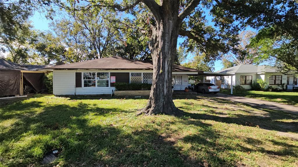 This photo showcases a single-story home with a large front yard and a mature tree. The house features a covered carport and a welcoming porch. It is situated in a neighborhood with similar-style homes and ample greenery.