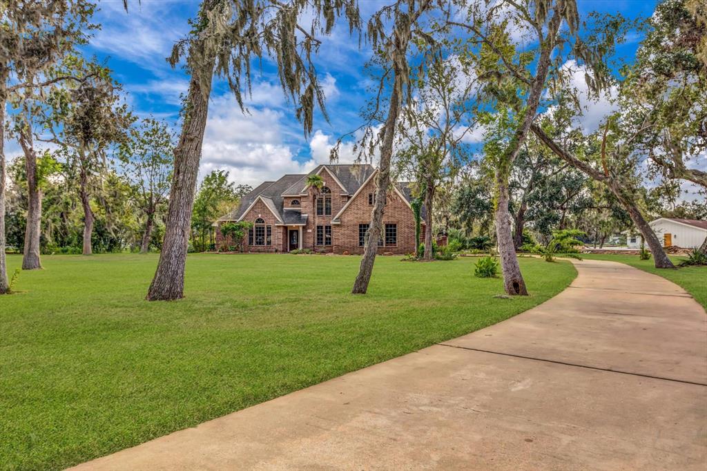 This is a spacious two-story brick home with a large, well-manicured lawn, mature trees, and a winding driveway leading to an attached garage. The property offers a tranquil, natural setting with ample outdoor space.