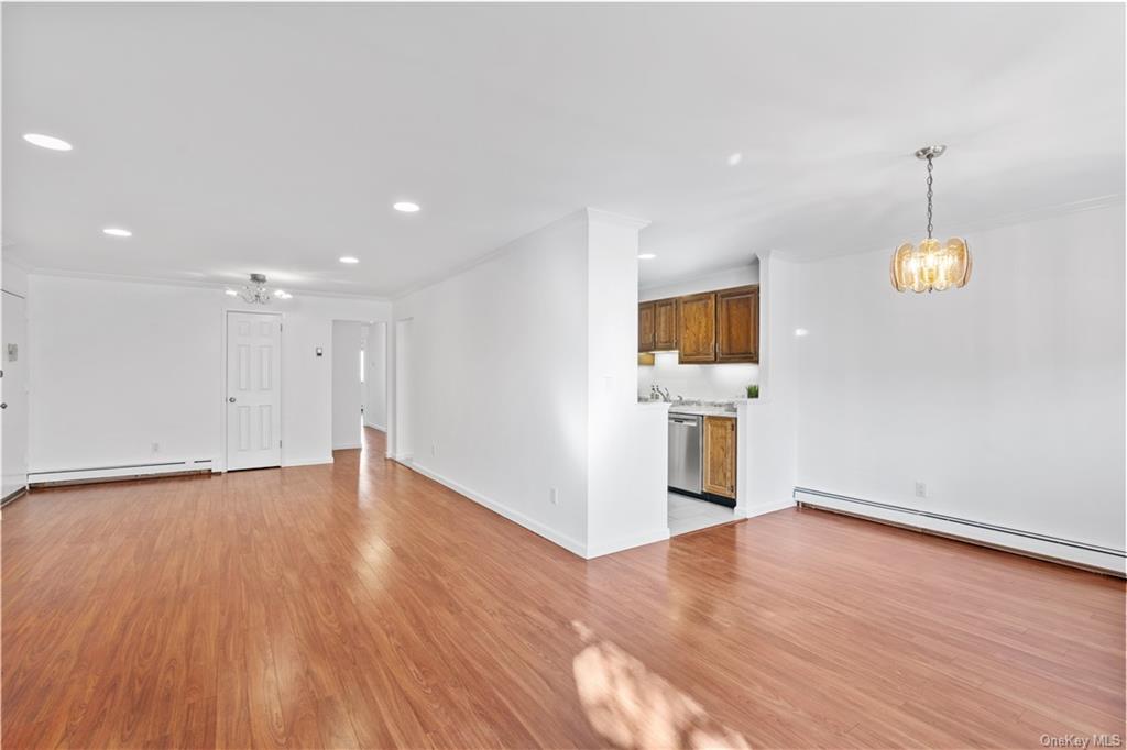 Living room open towards dining area and kitchen; featuring a chandelier, crown molding, medium brown wood-type flooring, and baseboard heating.