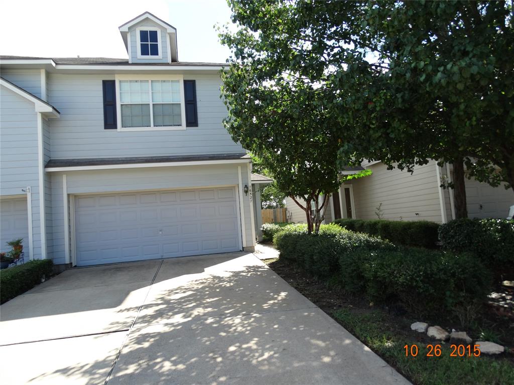 This is a two-story home featuring a single-car garage, a tidy front yard with shrubbery, and a tree providing shade. The house has a neutral color palette and a traditional design.