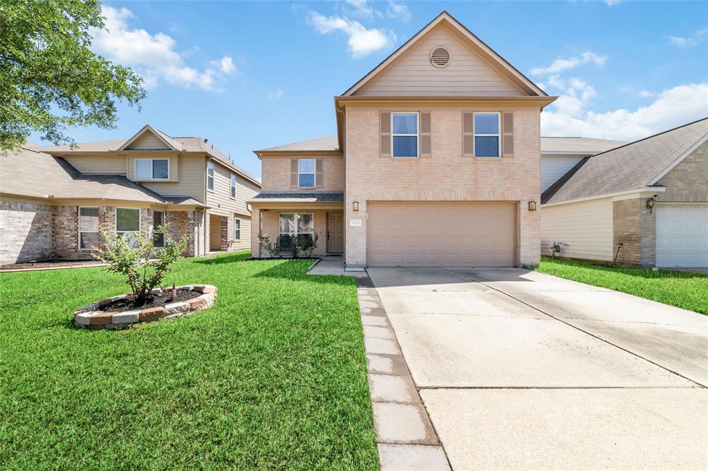 This is a two-story home with a light brick facade, featuring a two-car garage and a neatly maintained lawn. A small landscaped area adds a touch of greenery to the front yard.