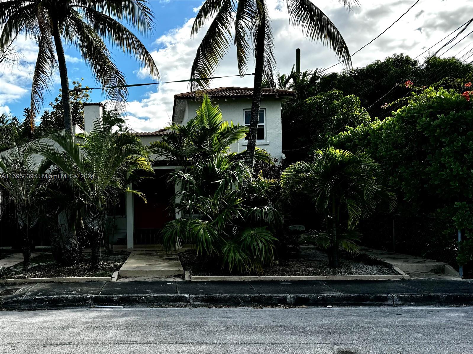 The image shows the entire front structure of the fourplex, which houses Units A, B, and C, while the cottage "Unit D", located in the back, is accessible via the narrow concrete pathway on the right partially obscured by tropical vegetation, including palm trees and lush shrubs.